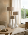 Set of three white sculptural floor lamps with natural linen drum shades, styled in a serene neutral-toned living room with curved sofas and timber accents