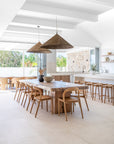 A beautifully styled dining space featuring a solid wood dining table with natural timber chairs, complemented by woven pendant lights and an open-plan kitchen with rustic stone accents