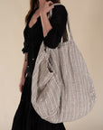 A woman in a black bohemian dress carrying a generously sized striped linen market bag with a relaxed, slouchy design and exterior pocket. The neutral-toned fabric and soft texture create a timeless, effortless style