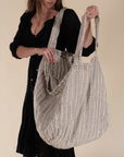 A woman in a black bohemian dress carrying a generously sized striped linen market bag with a relaxed, slouchy design and exterior pocket. The neutral-toned fabric and soft texture create a timeless, effortless style