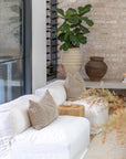 White linen sofa with neutral cushions styled in a serene living space featuring rustic ceramic pots, indoor plants, and a raw brick feature wall.