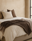 Sutu cotton throw in warm neutral and brown tones styled on a bed in a soft Mediterranean-inspired bedroom with natural light and rendered walls.