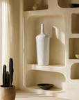 Tall white ceramic vase with a slender neck displayed on a sculptural built-in shelf, styled with earthy bowls and natural light.