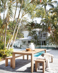 Teak carved chopping board styled on a solid timber outdoor dining table beside a pool, surrounded by lush palm trees and a relaxed coastal setting.