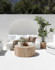 The Kulala Living Teak Drum Coffee Table styled in a bright outdoor setting. Crafted from natural teak branches, this round coffee table sits between two white lounge chairs with textured cushions, complemented by lush greenery and earthy decor for a serene alfresco retreat