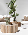 The Kulala Living Teak Drum Coffee Table styled in a bright outdoor setting. Crafted from natural teak branches, this round coffee table sits between two white lounge chairs with textured cushions, complemented by lush greenery and earthy decor for a serene alfresco retreat