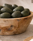 Large natural teak serving bowl filled with avocados, styled on a stone kitchen bench with neutral ceramics and olive oil in the background.