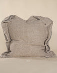 A textured linen cushion in a natural beige tone, featuring a relaxed, oversized design with raw-edge detailing, set against a neutral backdrop
