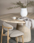 A beautifully styled dining space featuring a round travertine dining table, a neutral upholstered dining chair with timber accents, and a white ceramic vase with dried foliage. A ceramic cup, stacked plates, and a soft linen napkin add a touch of understated elegance
