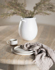 A beautifully styled dining space featuring a round travertine dining table, a neutral upholstered dining chair with timber accents, and a white ceramic vase with dried foliage. A ceramic cup, stacked plates, and a soft linen napkin add a touch of understated elegance