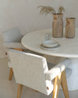 A serene dining space featuring a round travertine table with soft-textured upholstered chairs in a neutral palette. Wooden vases with dried foliage and minimalist ceramic tableware add organic warmth to the setting