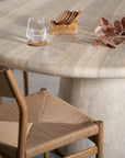 A beautifully styled travertine dining table featuring a wooden coaster set, a glass of water, and natural dried foliage, complemented by a rattan woven dining chair