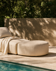 Travis Sofa and ottoman upholstered in natural fabric, styled with a throw blanket beside a swimming pool, creating a serene outdoor retreat