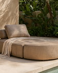 Travis Ottoman in latte fabric styled with a cushion and throw, placed beside a pool in an outdoor lounge setting with tropical greenery