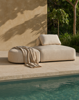 Travis Ottoman in natural fabric styled with a throw blanket, positioned poolside in an outdoor setting with stone tiles and greenery