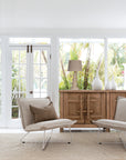Bright and airy living space by KULALA Living, featuring neutral-toned lounge chairs, a rustic wooden sideboard, and textured décor with lush garden views