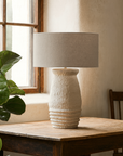 Udu Table Lamp with textured papier-mache base and linen shade styled on a timber table beside a window in a natural light-filled room