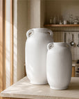 Pair of tall white amphora-style vases with twin handles, displayed on a stone kitchen bench in a light-filled, natural-toned kitchen.