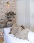 White linen sofa styled with textured neutral cushions, vintage ceramic pots, and stacked firewood against a rustic brick wall