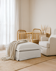 White linen ottoman with cushion and knitted throw in a neutral nursery, featuring a rattan cot, armchair, and warm natural decor.