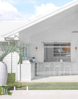 White coastal-style outdoor kitchen with a marble bar, white teak bar stools, brass tapware, and breezy open-plan design overlooking a manicured lawn.
