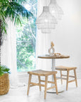 A serene dining nook featuring two white rattan pendant lights, a rustic round wooden table with a white pedestal base, and two natural wood stools with woven seats. Soft natural light filters through sheer curtains, enhancing the tranquil and airy aesthetic