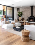 Light-filled living room featuring a white slipcover lounge, rustic stools, black timber coffee table, and a fireplace set against a natural brick wall, styled with earthy accents and neutral tones.