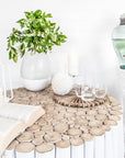 A beautifully styled setting featuring the Kulala Living White Teak Drum Coffee Table, adorned with an open book, candles, glassware, and a fresh greenery arrangement in a ceramic vase. The serene aesthetic is complemented by a glass water dispenser on a carved pedestal, creating a relaxed and inviting ambiance