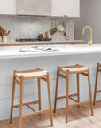 Close-up of KULALA Living teak barstools with woven seats, styled in a light-filled kitchen with white island bench, brass tapware, and artisanal tile splashback.