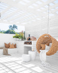 Natural wicker hanging egg chair with white cushion and throw, styled in a white outdoor patio space with built-in seating, firewood storage, and a pizza oven