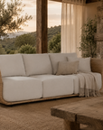 Malusi Sofa with a woven rattan frame and natural upholstered cushions styled in a warm resort-inspired living room beside a stone fireplace with flowing linen curtains and countryside views