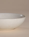 A handmade ceramic bowl with a smooth, speckled neutral glaze, set against a soft beige background. The minimalist design and organic shape make it a versatile piece for modern dining and home decor