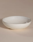 A handmade ceramic bowl with a smooth, speckled neutral glaze, set against a soft beige background. The minimalist design and organic shape make it a versatile piece for modern dining and home decor