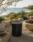 Black magnesium oxide stools arranged around an outdoor fire pit in a coastal garden retreat, creating a relaxed gathering space framed by native greenery and ocean views.