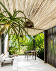 Tropical outdoor dining area under a eucalyptus pole pergola, featuring lush greenery, a rustic wooden dining table with metal chairs, and hanging pendant lighting. The space exudes a relaxed, resort-style ambiance