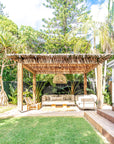 Tranquil outdoor pavilion featuring a natural timber pergola, latte pole roof, rattan pendant light, teak outdoor sofa, and tropical garden setting with lush greenery.
