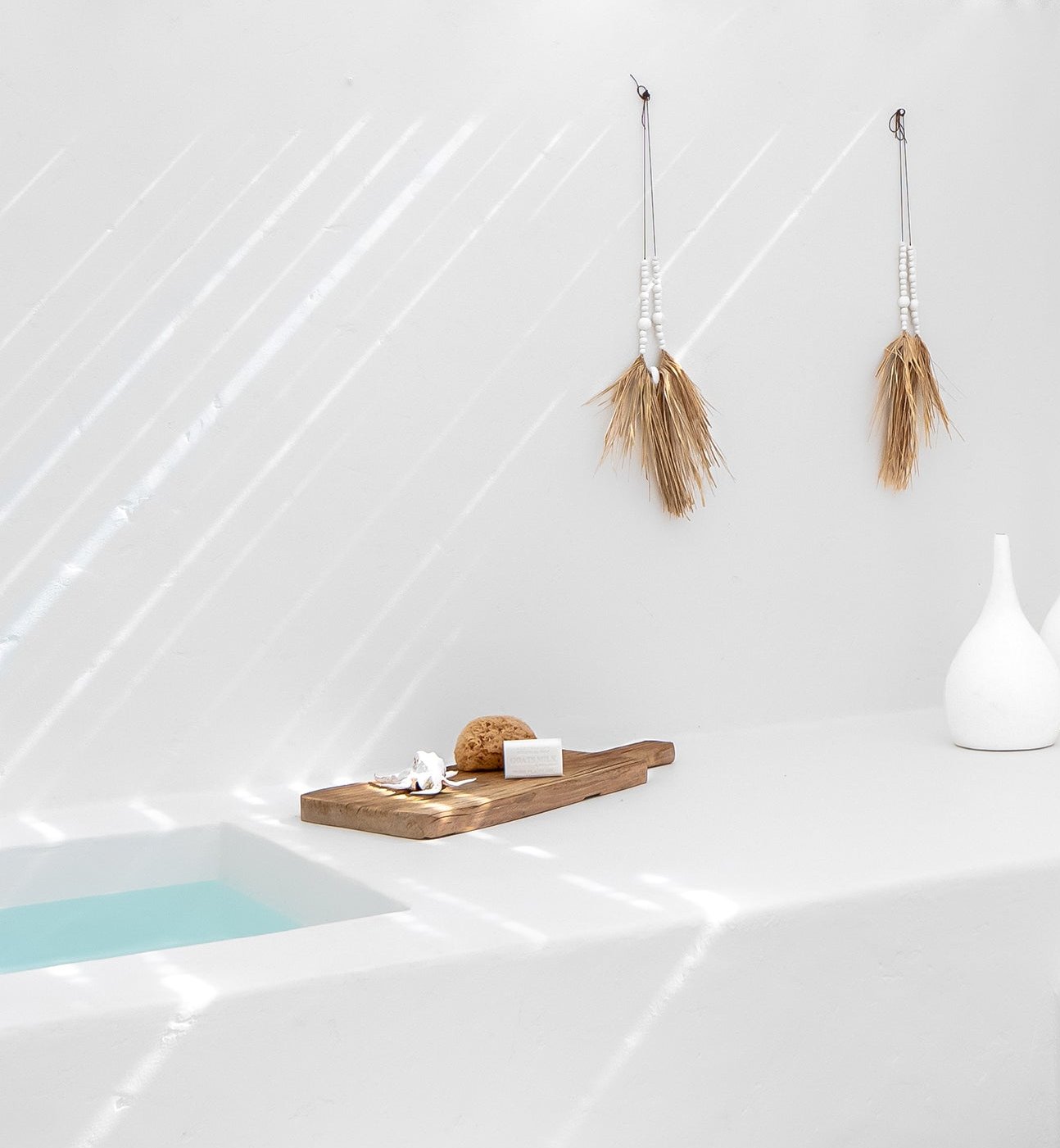 KULALA Living minimalist white bathroom featuring natural raffia and shell hanging decor, a wooden bath tray with soap and sponge, and a tranquil built-in bath