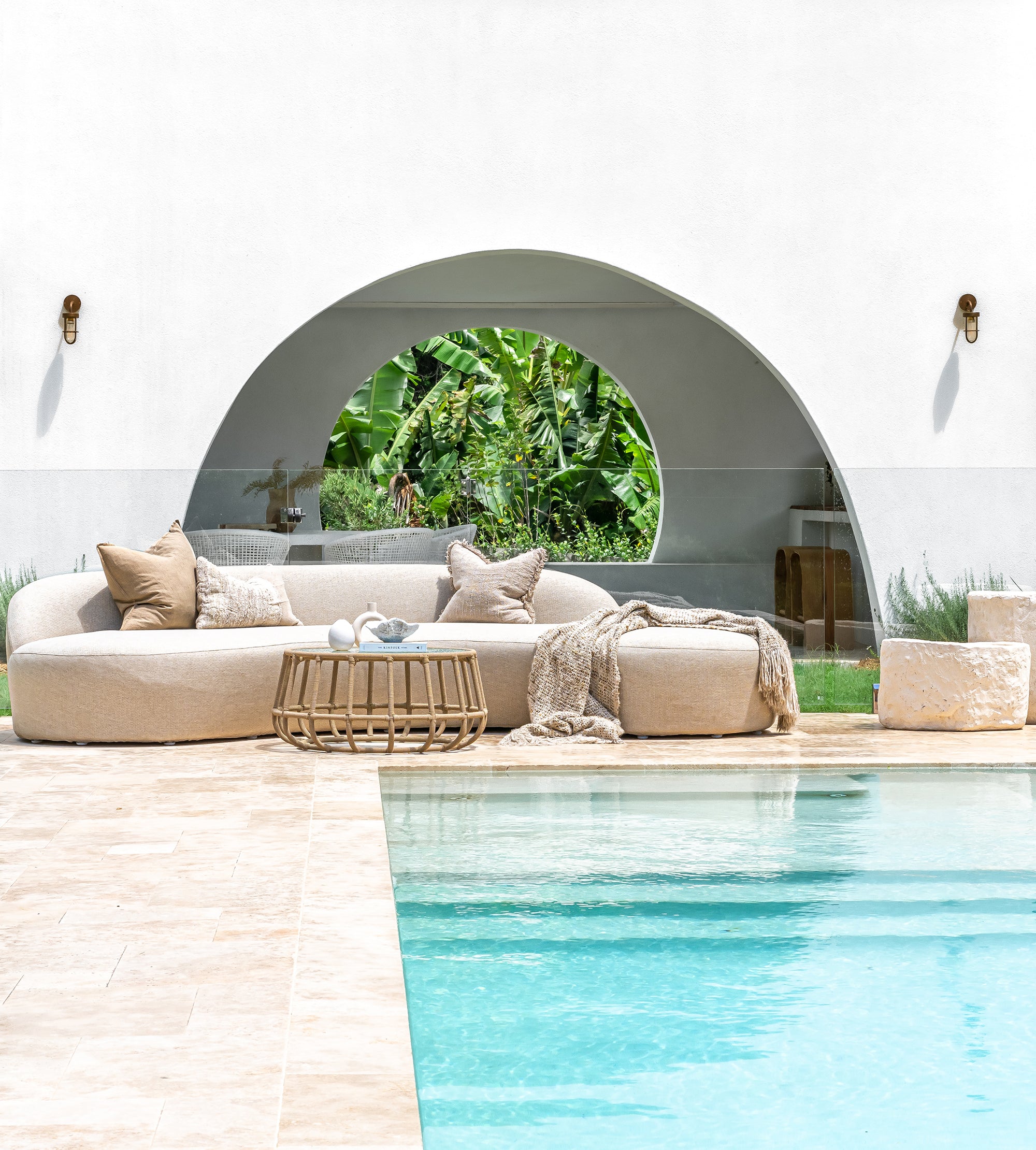 Beige outdoor sofa with cushions by a pool, featuring a circular mirror reflecting greenery.