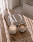 A styled interior scene with a lit white candle and a cup of latte on a rustic wooden table beside a book, in soft natural light with window shadows.
