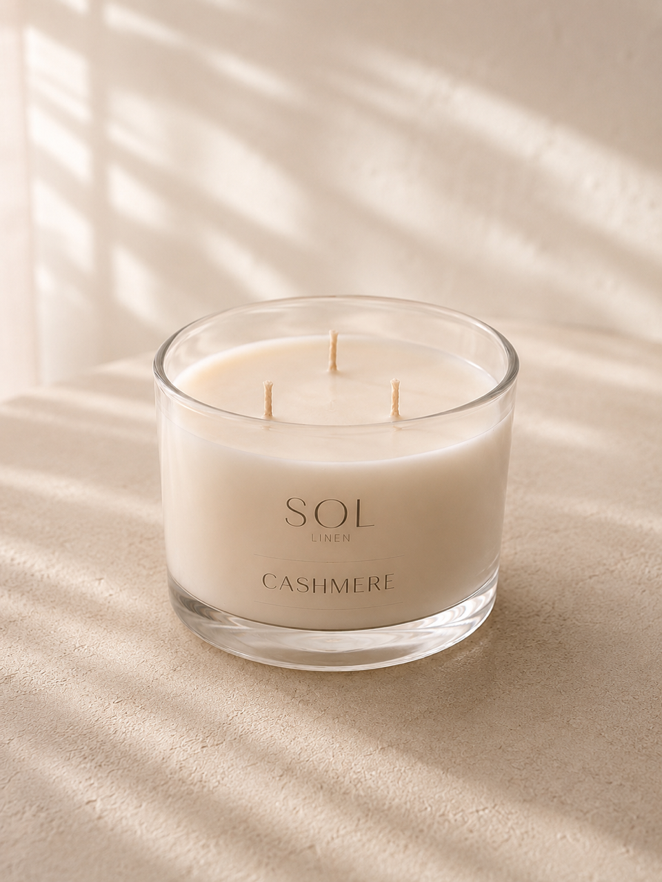 A three-wick soy candle in a clear glass jar with cream-colored wax, labeled 'SOL CASHMERE', set on a textured beige surface with soft window shadow patterns.