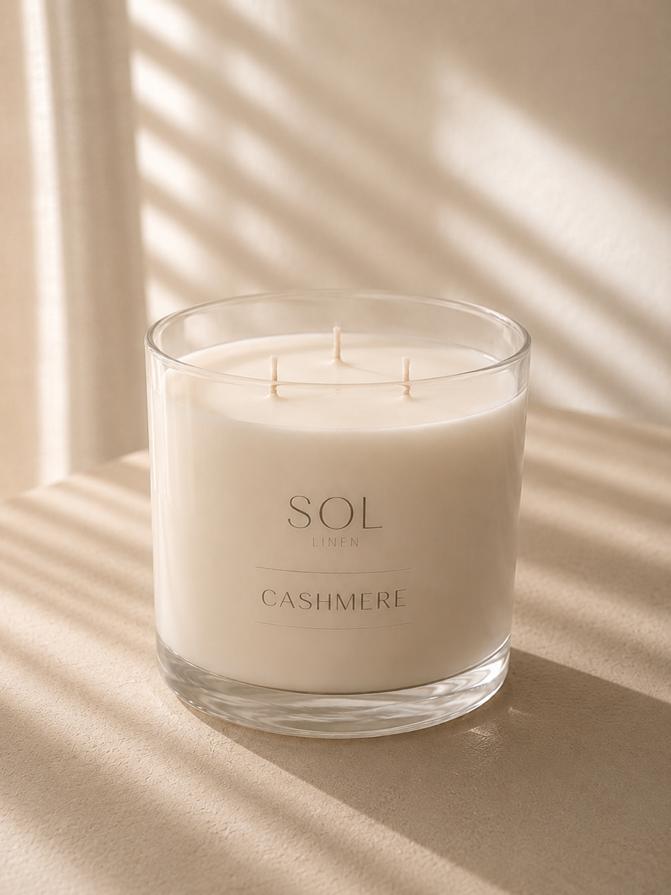 A three-wick soy candle in a clear glass jar with cream-colored wax, labeled 'SOL LIGHTS CASHMERE', set on a beige surface with soft window shadow patterns.