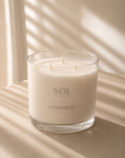 A three-wick soy candle in a clear glass jar with cream-colored wax, labeled 'SOL LIGHTS CASHMERE', set on a beige surface with soft window shadow patterns.