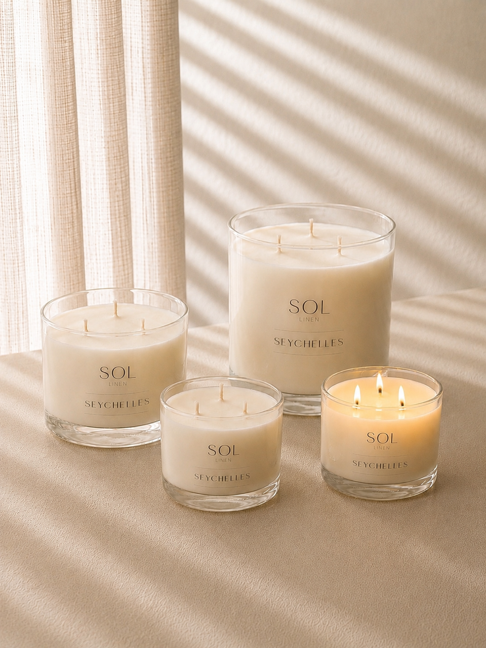 Four SOL by Seychelles candles in clear glass jars, with three unlit and one lit, arranged on a beige fabric surface with soft window shadow patterns.