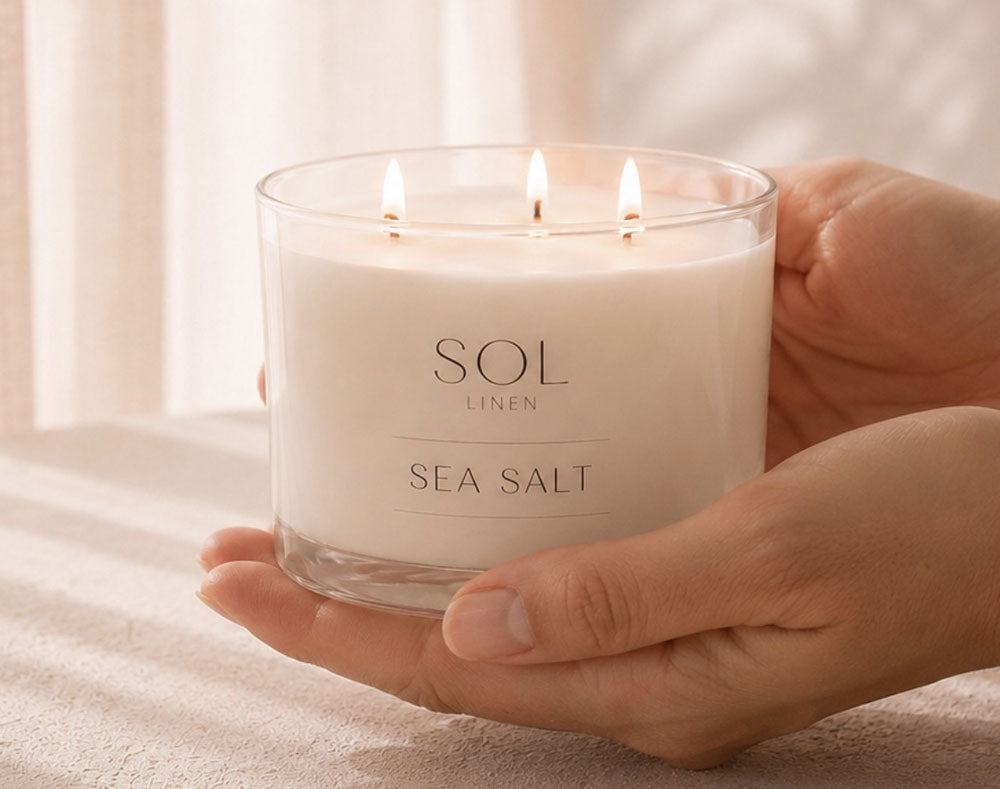 A person's hands holding a lit three-wick candle in a clear glass jar. The candle is labeled 'SOL LINEN SEA SALT' and has creamy off-white wax.