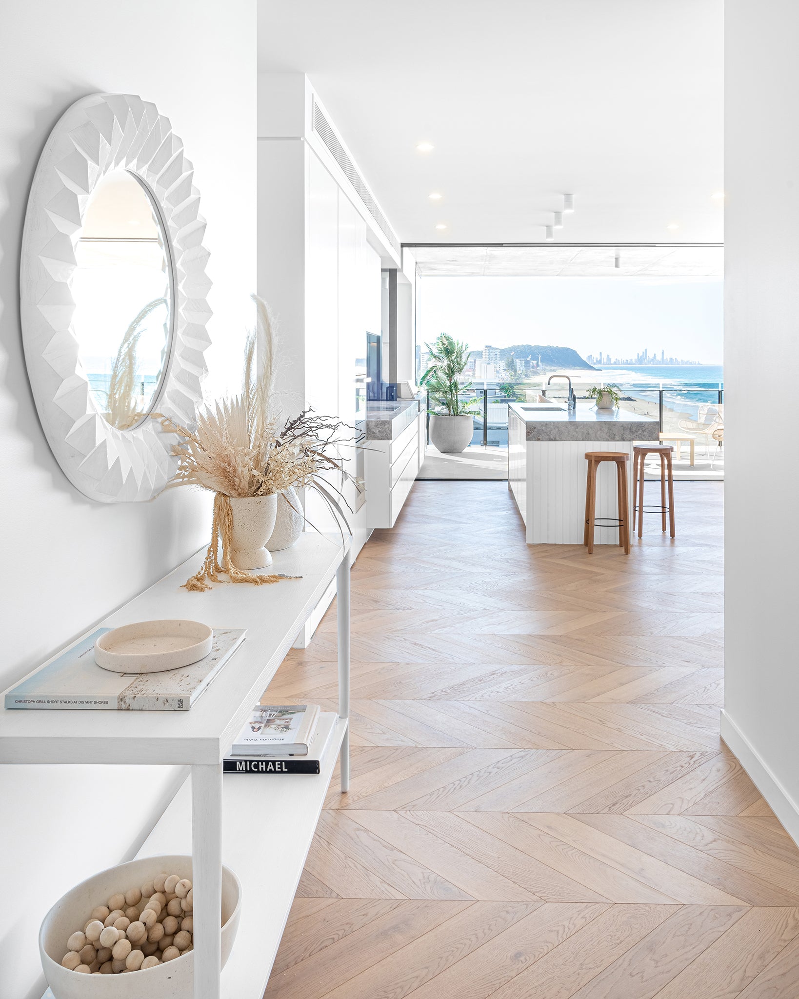 A bright and airy coastal home featuring herringbone oak flooring, a sculptural white mirror, and Kulala Living solid oak bar stools at a marble island. Floor-to-ceiling glass doors open to an ocean-view balcony, blending indoor and outdoor living seamlessly