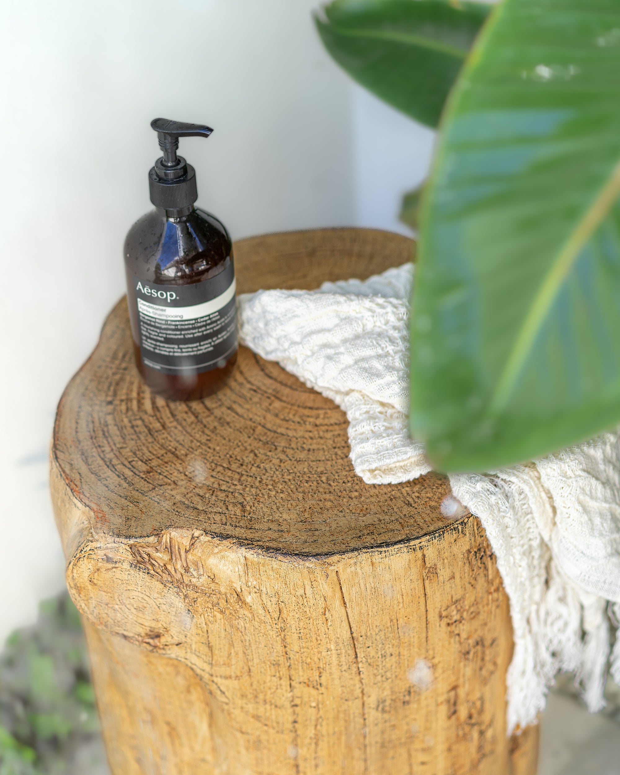 Aesop hand soap resting on a rustic timber stump side table beside a textured white linen hand towel, surrounded by lush green leaves in an outdoor bathroom setting.