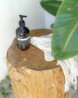 Aesop hand soap resting on a rustic timber stump side table beside a textured white linen hand towel, surrounded by lush green leaves in an outdoor bathroom setting.