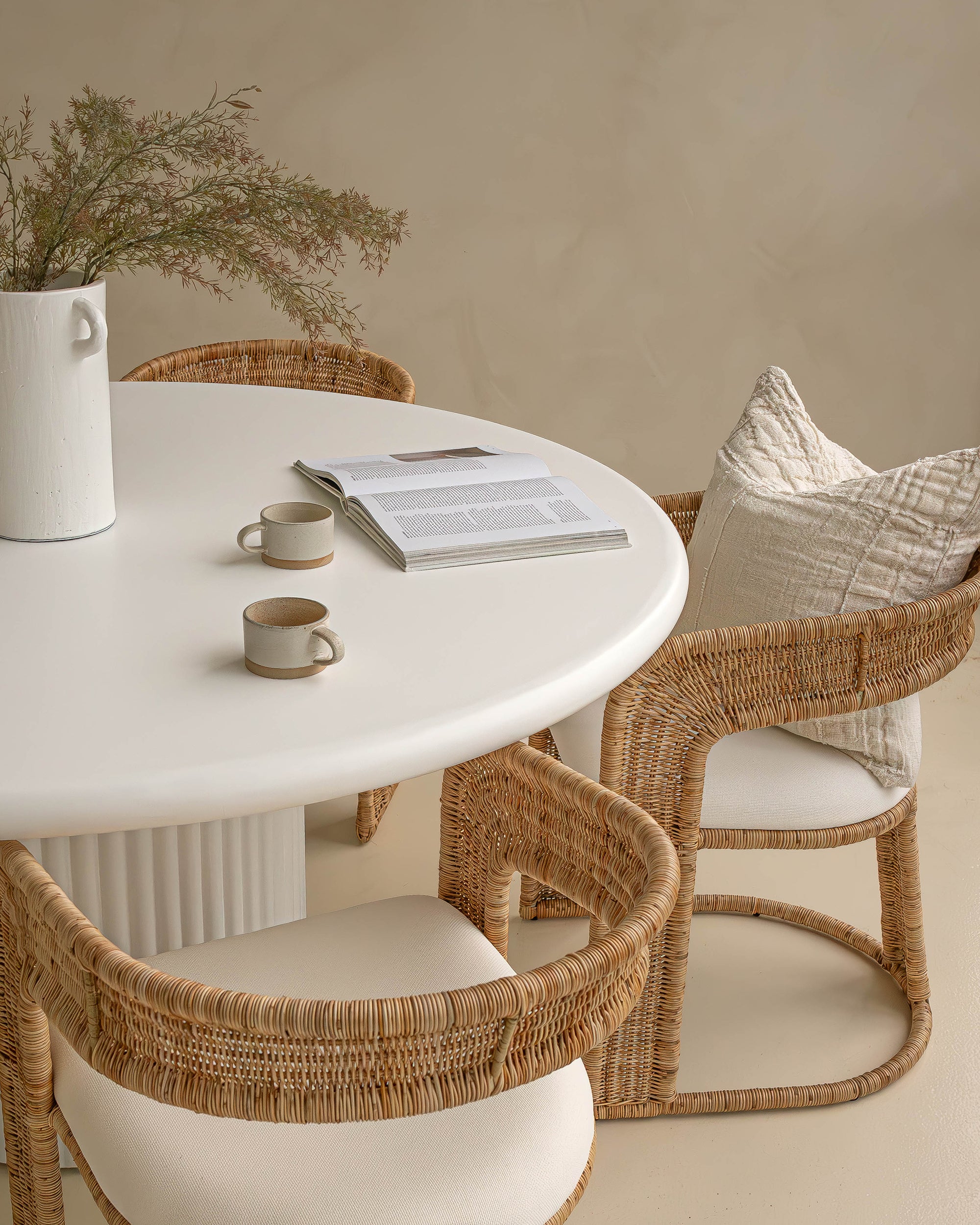 Coastal-inspired dining space featuring curved rattan dining chairs with upholstered seats, paired with a white fluted dining table. Styled with a ceramic vase, neutral-toned decor, organic ambiance