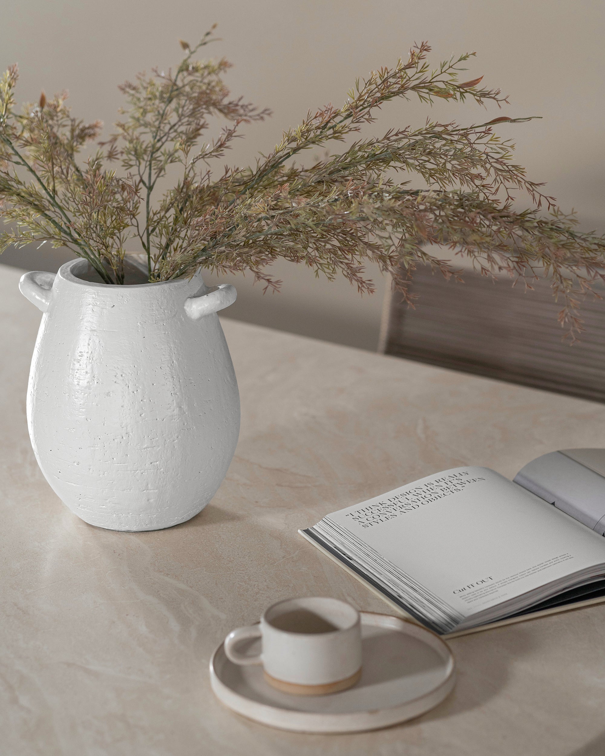 Textured white ceramic vase with delicate greenery, styled on a hydro-dipped concrete table alongside an open book and a handmade ceramic coffee cup. A serene and minimalist setting with organic textures and neutral tones