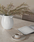 Textured white ceramic vase with delicate greenery, styled on a hydro-dipped concrete table alongside an open book and a handmade ceramic coffee cup. A serene and minimalist setting with organic textures and neutral tones